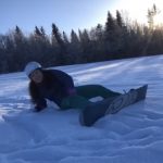 Jente har akkurat falt på snowboard og ligger i snøen og ler. I bakgrunnen er det trær og fin sol og blå himmel.