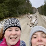 Selfie tatt av to blide jenter på tur med litt snø rundt og to reinsdyr som går bak dem.