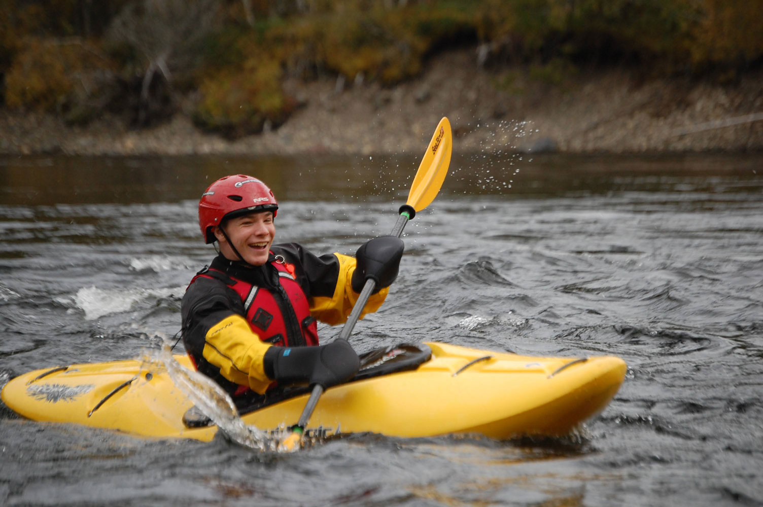 Elev i rød hjelm i gul elvekajakk smiler og padler i elva.