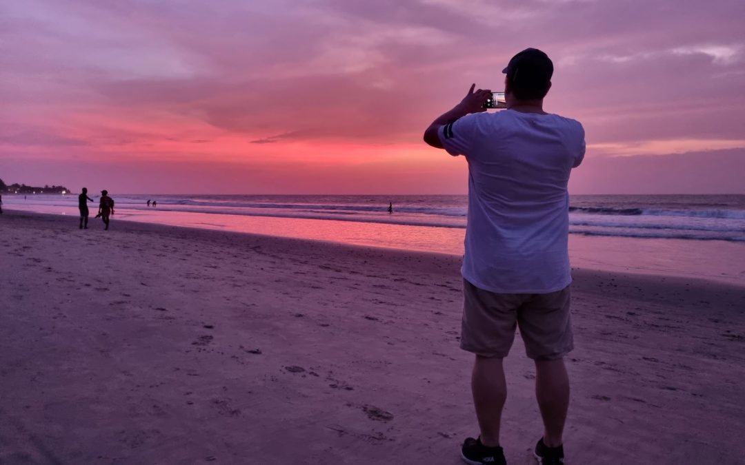 Mann står og tar bilde av solnedgangen på stranda.