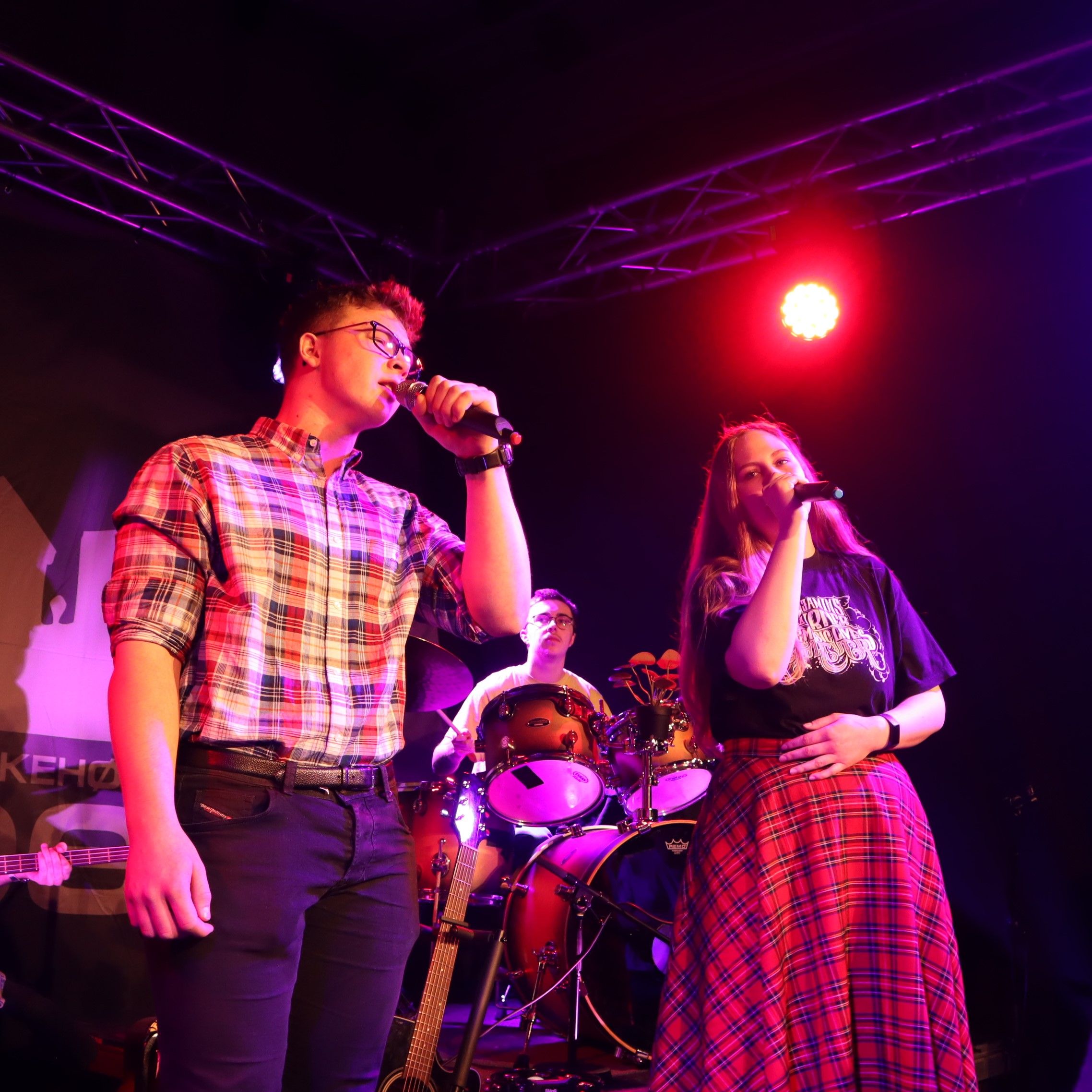 To personer står på scenen på Vefsn Folkehøgskole Toppen og synger duett. I bakgrunnen ses en person som spiller trommer.