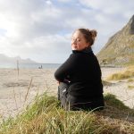 Johanne Taraldsen sitter på gresset i solen med ryggen til, snur seg bakover og smiler til fotografen. I bakgrunnen en strand i Lofoten.
