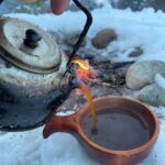 Hånd holder en kaffekjele som heller kaffe i ei kokse. I bakgrunnen bålet. Vinter.