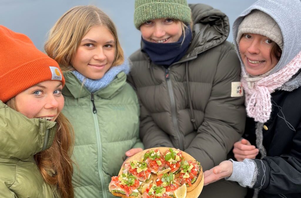 Kokkekamp ute på Vefsn Folkehøgskole Toppen. Fire elever viser fram maten de har laget. De har på seg dynejakker og lue. Vefsnfjorden i bakgrunnen.