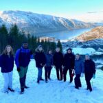 Gruppe elever fra Vefsn Folkehøgskole Toppen poserer på tur til Tårnet. Vinterkledd og med rumpeakebrett i hånda. Snø på bakken. I bakgrunnen Halsøutoppen og Vefsnfjorden.